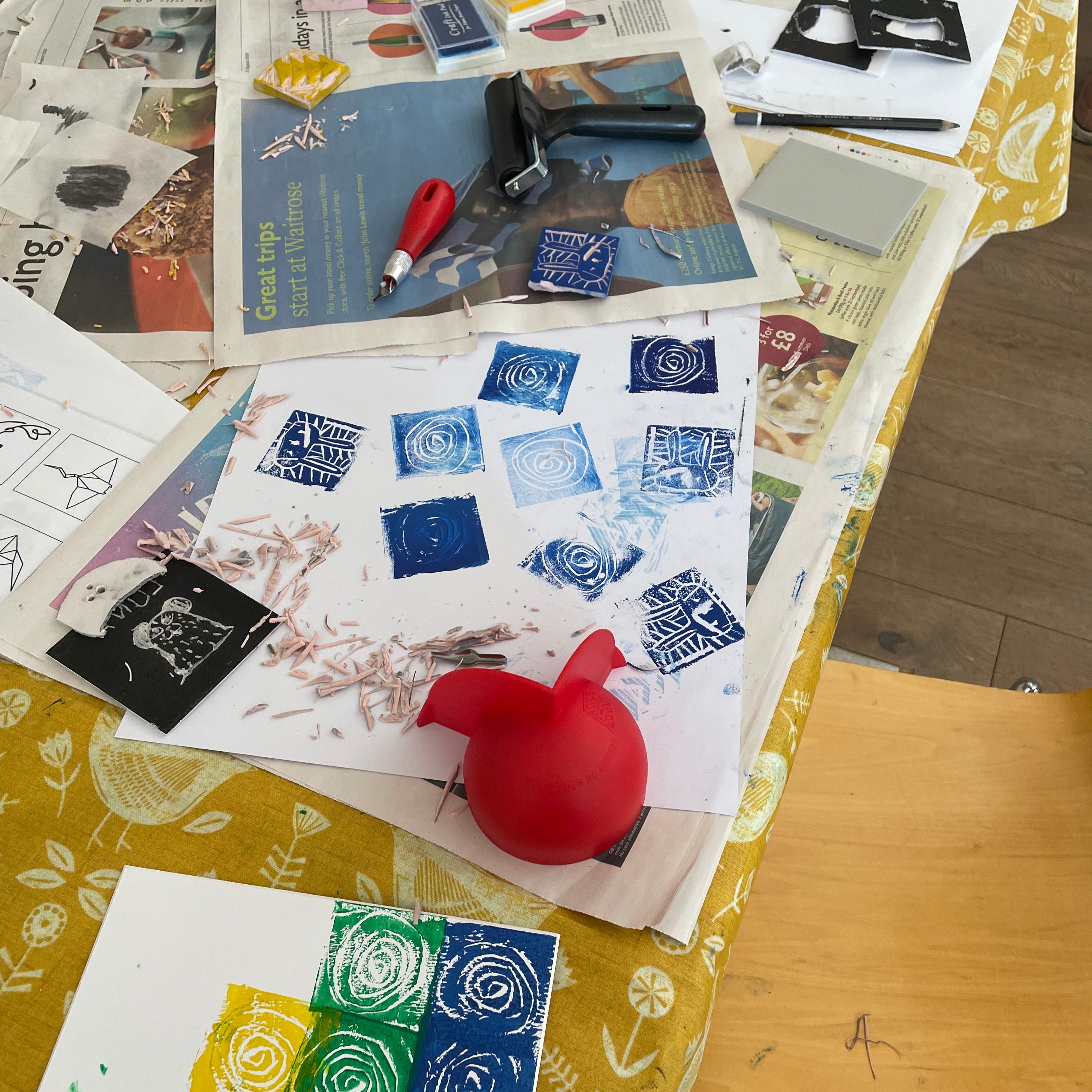 A tabletop covered with art and craft supplies, including blue and black rubber stamps, printed papers, a red stamping tool, and a yellow-and-white floral tablecloth. No person is visible, but the real indoor environment and surrounding furniture make this Lifestyle rather than Product/Packaging.