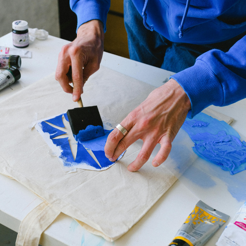 The image shows an adults hands stencilling a print onto a white tote bag